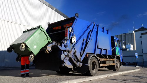 A large blue commercial rubbish collection truck with a hydraulic lift system at the rear is actively collecting waste from a green wheeled bin that is being hoisted into the truck's container. The green bin is made of plastic and features a textured surface with a black lid, situated on a paved surface beside a white industrial building. A worker wearing high-visibility red trousers and reflective safety gear stands nearby, guiding the bin's placement. The background shows a bright blue sky with some clouds, and the truck is positioned in a parking area or service yard, likely in Oxted or nearby within Surrey. The scene reflects typical urban rubbish removal operations, emphasizing waste collection and disposal services provided by Commercial Waste Oxted, with a focus on waste management and rubbish removal in the local area.