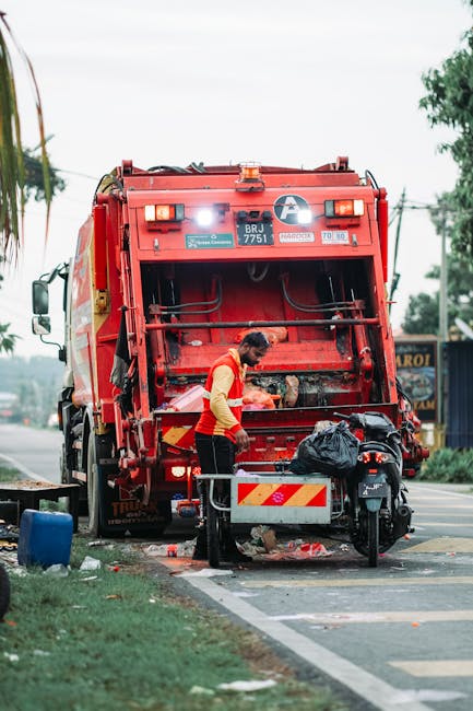 A large pile of mixed waste materials situated on a paved driveway outside a commercial property. The waste includes black plastic bins, cardboard boxes, and discarded packaging, with some items stack