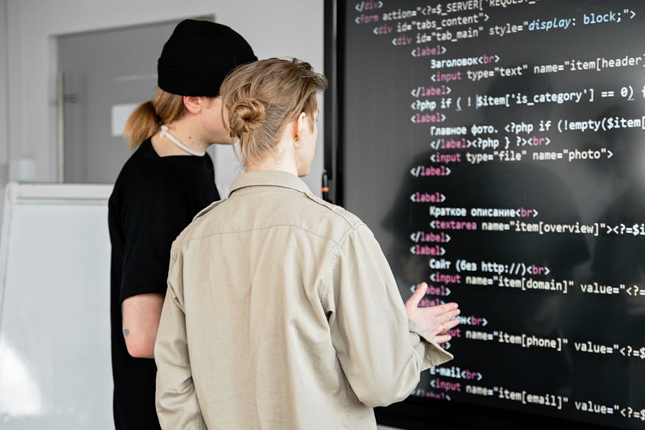 Two women stand in front of a large digital screen displaying lines of code, with one woman pointing at the screen while the other observes. The woman in the foreground has light brown hair tied in a