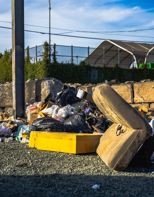 A close-up view of a pile of mixed rubbish and waste materials on a gravel surface in an outdoor setting. The pile includes black plastic garbage bags, a yellow plastic container, an old, discolored c